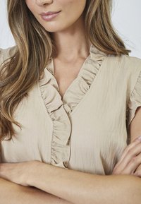 Beige Bluse mit Rüschenausschnitt, kurzen Ärmeln und weicher Textur. Die Kleidung ist durchgeknöpft und hat einen lockeren Schnitt, geeignet für lässige Anlässe.