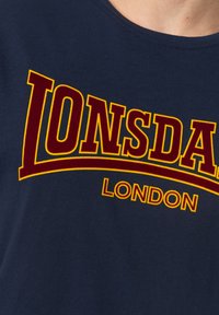 Tums tumši zils kokvilnas T-krekls ar lielu bordo krāsas "LONSDALE" uzrakstu un dzeltenu kontūru, apaļa kakla izgriezuma, ar mīkstu tekstūru.