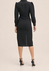 Schwarzes, tailliertes Kleid mit einem Kragen, langen Puffärmeln und einer Länge bis zur Wade. Verfügt über einen Schlitz hinten und einen strukturierten Stoff. Kombiniert mit schwarzen High Heels.