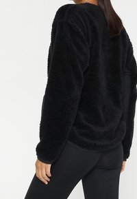 Zwarte, pluizige trui met een losse pasvorm, voorzien van een ronde hals en lange mouwen, getoond vanaf de achterkant tegen een effen achtergrond.
