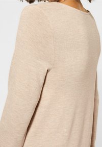 Beige neulepaita, tehty pehmeästä, ribatusta kankaasta. Siinä on pyöreä kaula-aukko ja pitkät hihat, joissa näkyy sauman yksityiskohdat olkapään ja käden alueella.