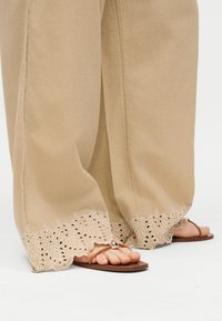 Μπεζ παντελόνι φαρδιάς γραμμής με κεντημένες κυματιστές άκρες, συνδυασμένο με καφέ επίπεδα σανδάλια που έχουν μία χρυσή διακοσμητική χάντρα στα λουριά.