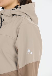 Beige and brown outdoor jacket featuring a hood, textured fabric, and a reflective logo on the sleeve. Adjustable drawstrings at the collar.