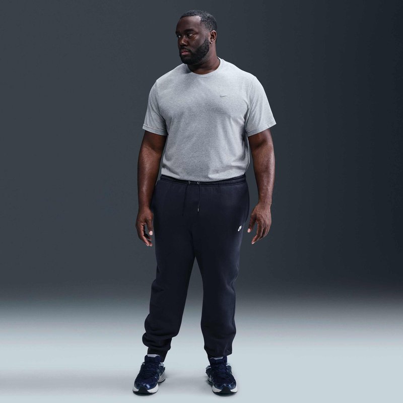 Uomo in piedi, indossa una t-shirt grigia a maniche corte, pantaloni jogger neri e scarpe da ginnastica nere, guarda alla sua sinistra su uno sfondo scuro e uniforme.