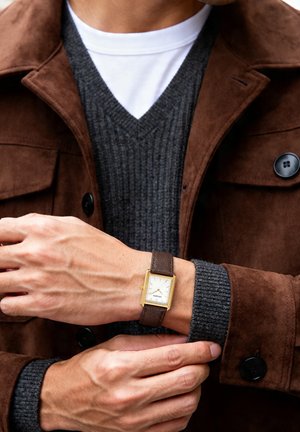 Mann stellt rechteckige goldene Uhr mit braunem Lederarmband ein, trägt braune Wildlederjacke und dunkelgrauen V-Ausschnitt-Pullover über weißem Hemd.