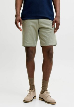 Mężczyzna ubrany w beżowe szorty, brązowe skarpetki, jasnobrązowe sznurowane buty i ciemnoniebieską koszulę na jednolitym jasnym tle.