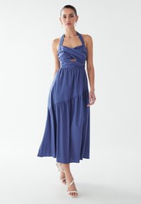 Žena nosi bezrameni plavi halter midi haljinu s ukriženim detaljima od tkanine i izrezom, uparenu s bijelim sandalama s trakama i bisernim naušnicama.