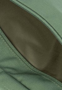 Grüner Stoff mit einer strukturierten Oberfläche, mit einer glatten, dunkelgrünen Fütterung, die durch einen Reißverschluss sichtbar ist. Genähte Kanten.