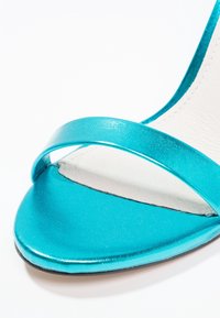 Gros plan d'une sandale turquoise métallique à bout ouvert avec une fine lanière sur les orteils et une couture de semelle intérieure blanche.