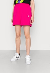 Bright pink cotton shorts with an elastic waistband and side seams, featuring a white Adidas logo. Paired with white sneakers.