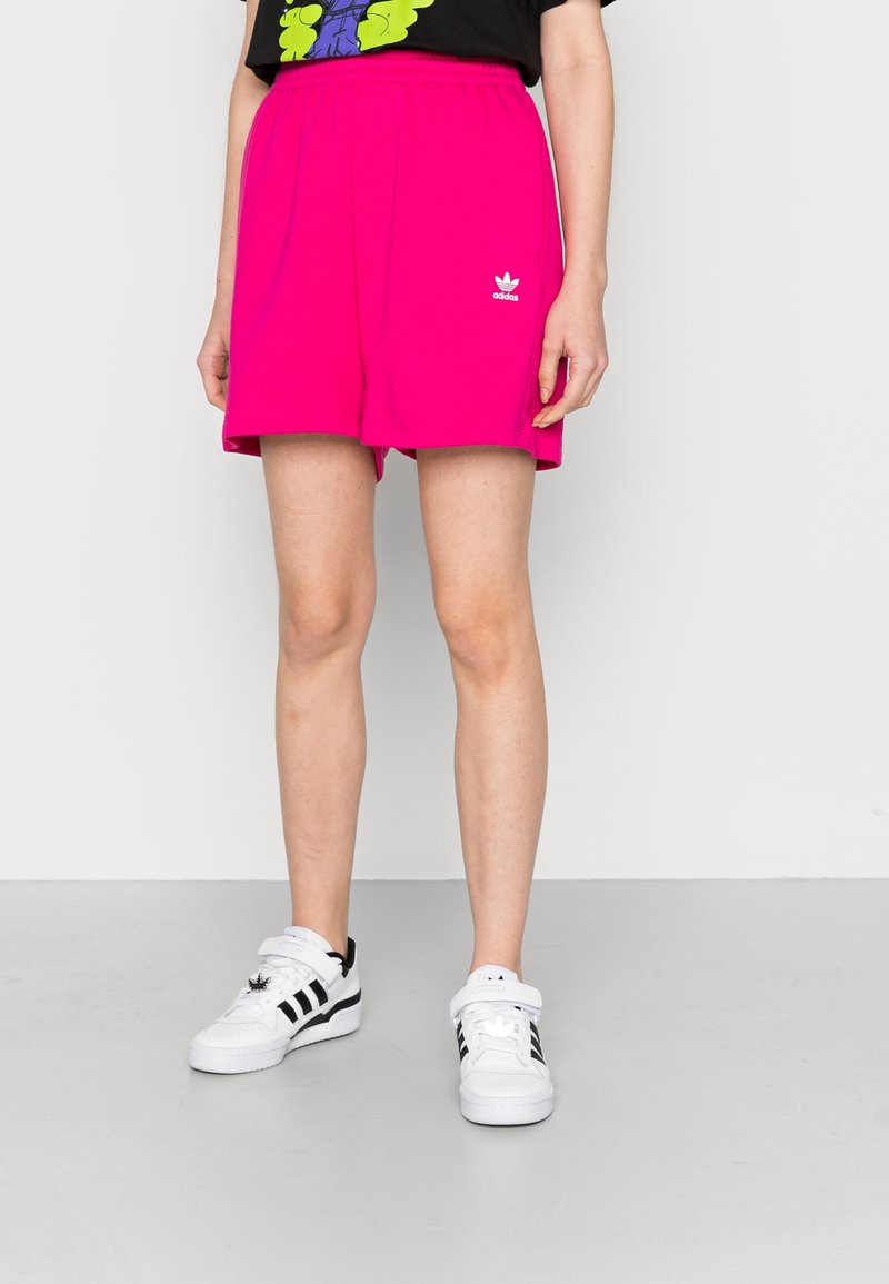 Bright pink cotton shorts with an elastic waistband and side seams, featuring a white Adidas logo. Paired with white sneakers.