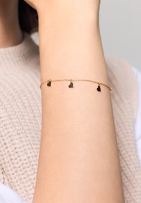 Złoty bransoletka łańcuszkowa z trzema małymi zawieszkami w kształcie serca, delikatnymi ogniwami i gładką, połyskującą powierzchnią na tle koloru skóry.