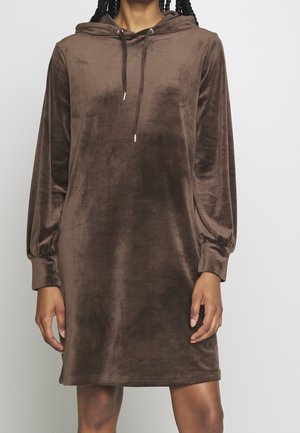 Femme portant une robe marron à capuche avec manches longues en tissu velouté doux, debout, les bras détendus le long du corps.