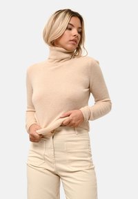Maglione beige a collo alto realizzato in materiale morbido, con una vestibilità aderente e polsini a costine, abbinato a pantaloni chiari a vita alta.