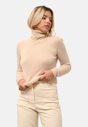 Beige Rollkragenpullover aus weichem Material, mit schlanker Passform und gerippten Bündchen, kombiniert mit hellen, hochgeschnittenen Hosen.