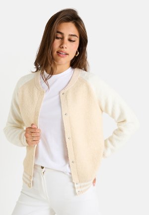 Cárdigan color crema con textura suave y peluda. Presenta un cierre de botones, cuello acanalado y puños a rayas en tonos más claros. Llevado sobre una camiseta blanca.