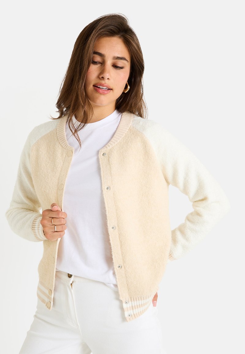 Cárdigan color crema con textura suave y peluda. Presenta un cierre de botones, cuello acanalado y puños a rayas en tonos más claros. Llevado sobre una camiseta blanca.