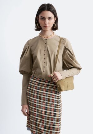 Vrouw draagt een beige blouse met pofmouwen, gouden sieraden, een geruite rok en draagt een kleine zandkleurige schoudertas, staand tegen een effen achtergrond.