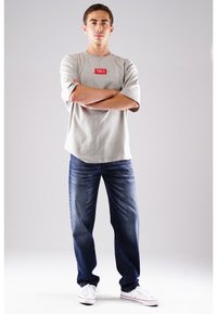 Graues Kurzarmhemd mit rotem Logo, kombiniert mit dunkelblauen Jeans und weißen Sneakern. Das Modell steht mit verschränkten Armen vor einem neutralen Hintergrund.