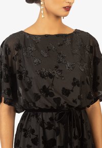Robe noire avec un tissu transparent et texturé orné d'appliqués floraux. Présente une ceinture à la taille et des manches larges et fluides.