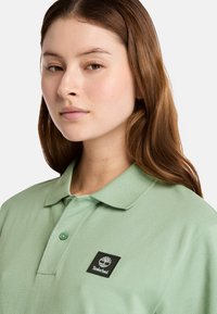 Ljusgrön pikétröja med texturerad tygkvalitet, prydd med en svart Timberland-logopatch på vänster sida och en klassisk kragsdesign.