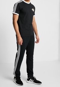 Svart bomullströja med vita accenter och logotyp, tillsammans med svarta träningsbyxor med vita sidostrålar. Atletiska skor syns också.