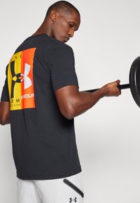 Crna atletska majica s velikim višebojnim logotipom na leđima. Model drži uteg i nosi svijetle hlače sa detaljem na zatvaraču.