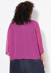 Kvinna med lockigt hår som bär en genomskinlig magentablus med trekvartsärmar och en mörk kjol, sedd bakifrån mot en vit vägg.