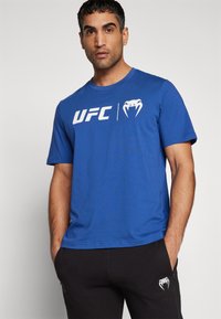 Blå bomulds t-shirt med hvid "UFC" logo og slange grafik. Korte ærmer, rund hals og afslappet pasform. Bæres med sorte sweatpants.