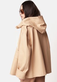 Beige übergroße Kapuzenjacke mit Knopfschluss und Kordelzug, breite Ärmel und eine strukturierte Stoffoberfläche.