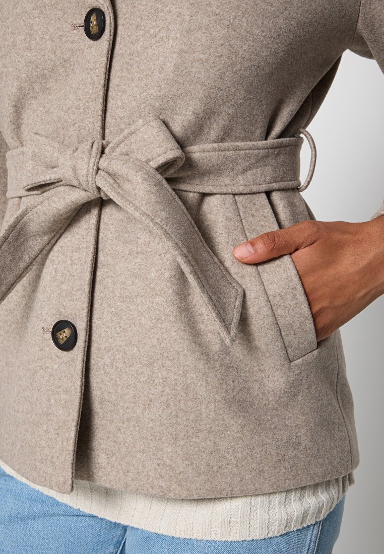 Cappotto in misto lana beige chiaro con vita aderente, grande bottone nero e tasche laterali, indossato sopra un maglione bianco a coste e jeans blu.