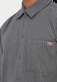 Szürke gombos ing fehér csíkokkal, gallérral, két gombbal az elülső részen, valamint egy bal oldali zsebbel, amelyen a Dickies logó található.