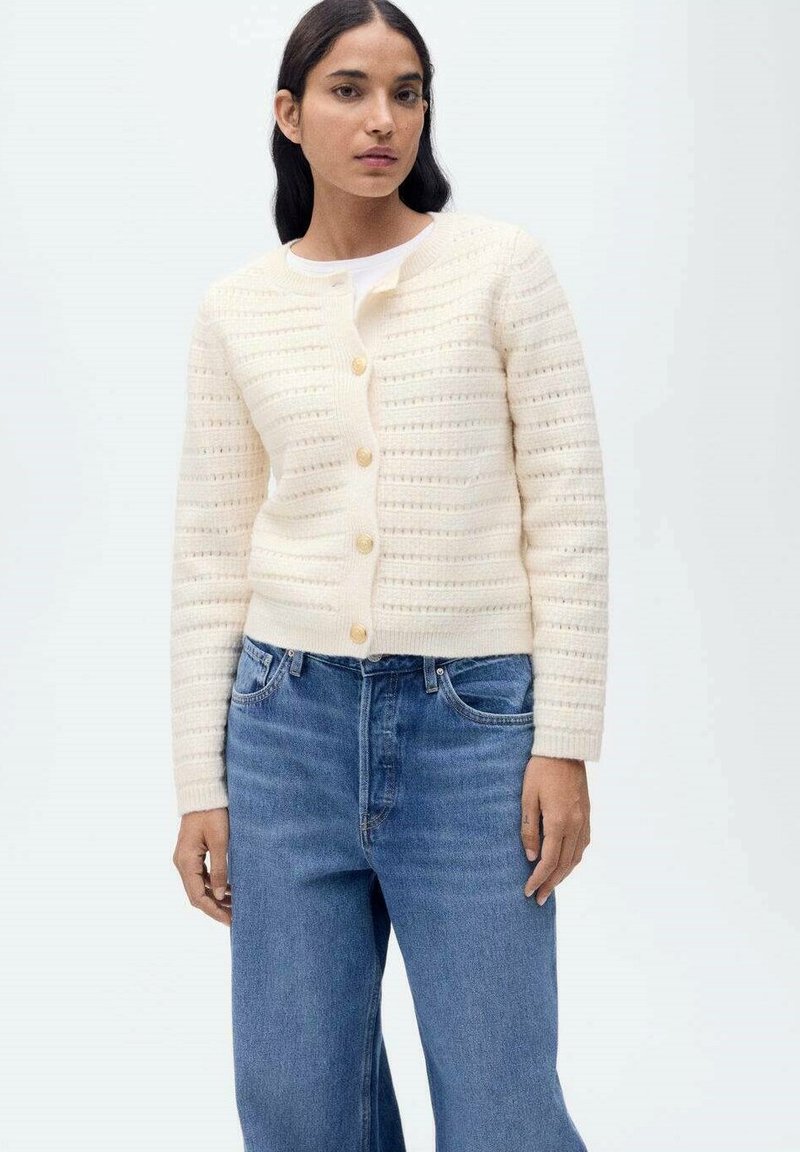 Strick-Cardigan in Cream mit geripptem Muster, Rundhalsausschnitt, Knopfverschluss und langen Ärmeln, kombiniert mit hellblauen Jeans.