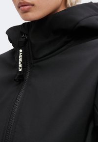 Gros plan d'une personne portant une veste noire avec un col haut et une tirette de fermeture éclair étiquetée "LEADER."
