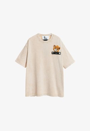 Camiseta de algodón beige con mangas cortas y un escote redondo, que presenta un bordado de un oso de peluche en el lado izquierdo del pecho y una etiqueta negra.