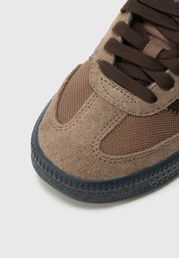 Brązowy zamszowy i siateczkowy sneaker z ciemnobrązowymi sznurówkami, teksturowaną gumową podeszwą oraz wyraźnymi detalami szwów. Okrągły czubek i niska sylwetka.