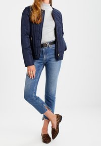 Marineblauwe quilted bomberjack over een grijze coltrui, gecombineerd met lichtblauwe cropped jeans en loafers met luipaardprint. Opvallende zijsplitten in de jeans.