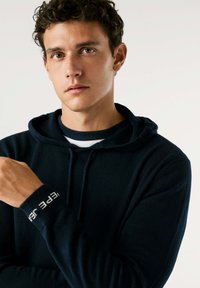 Model navy blu hoodies iz mehkega materiala, z vrvico, rebrastim manšetam in napisom "ohrani to resnično", ter sproščenim krojem.