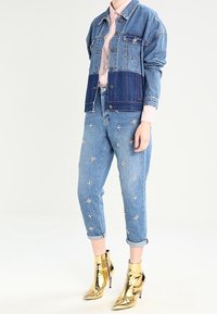 Denim jacket with light and dark blue patches, pink shirt, and distressed jeans featuring floral embroidery. Shiny gold ankle boots.