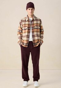 Overhemd van flanel in bruin-wit geruit, gecombineerd met bordeauxrode ribfluwelen broek. Accessoires omvatten een bordeauxrode gebreide muts en witte sneakers.