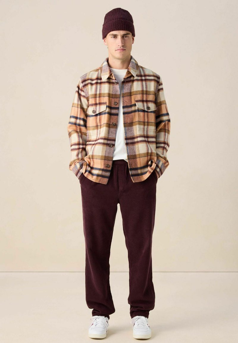 Overhemd van flanel in bruin-wit geruit, gecombineerd met bordeauxrode ribfluwelen broek. Accessoires omvatten een bordeauxrode gebreide muts en witte sneakers.