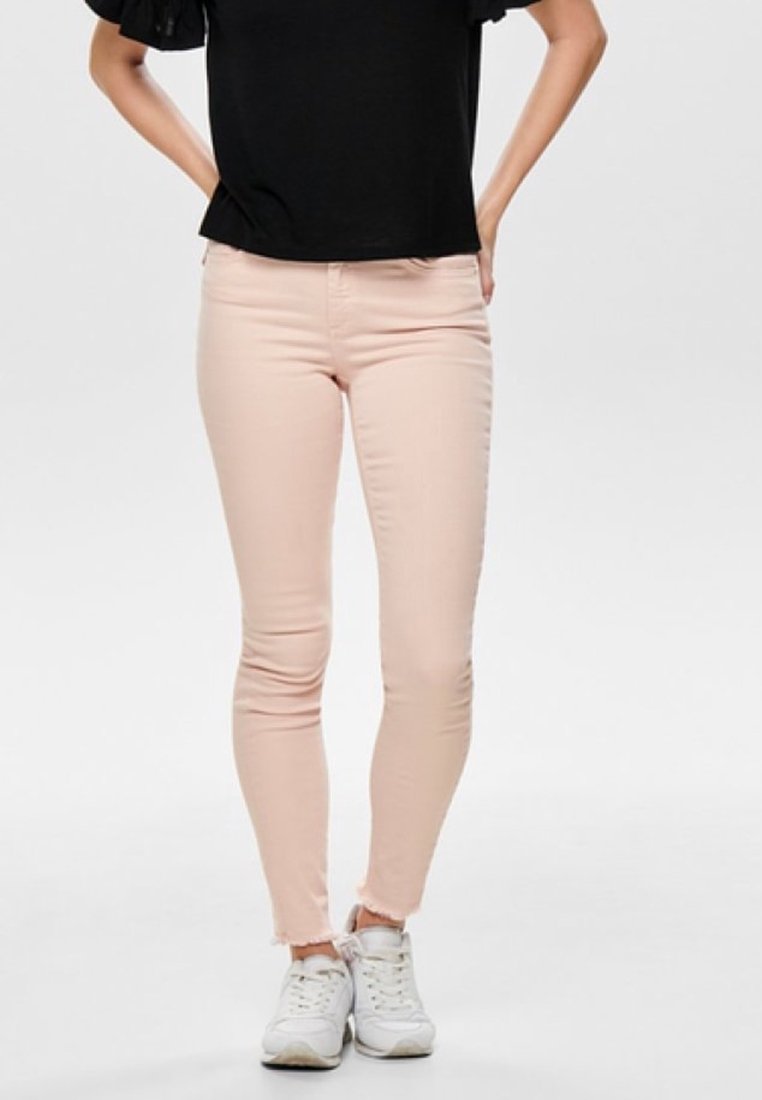 Jean skinny rose clair avec un ourlet effiloché, associé à un haut noir. Porté avec des baskets blanches, affichant un design décontracté et ajusté.
