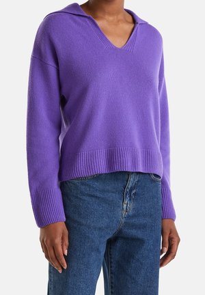 Personne portant un pull violet en maille à col en V avec capuche et un jean en denim bleu, debout avec les bras détendus contre un fond uni.