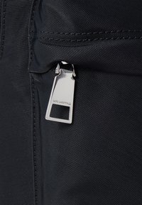 Tissu noir texturé avec une fermeture éclair argentée et des détails de tag. Le logo est gravé sur le tirette de la fermeture éclair, mettant en valeur la marque.