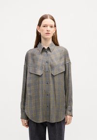 Camicia a quadri grigia in tessuto morbido, con due grandi tasche sul petto, chiusura con bottoni frontale e orlo curvo. Maniche lunghe con polsini abbottonati.