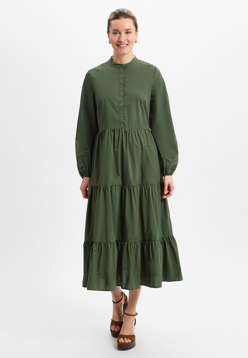 Langes grünes Baumwollkleid mit einem gestuften Rock, Knopfleiste vorne und langen Puffärmeln. Akzentuiert mit einem hohen Kragen und gerafftem Taillenteil.