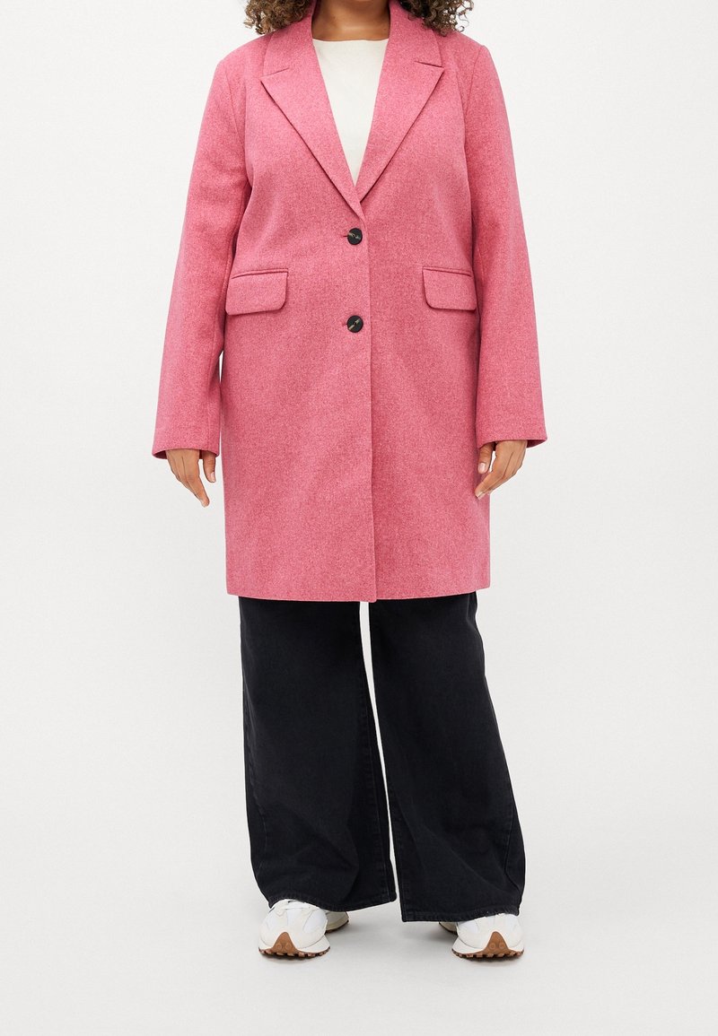 Abrigo de mezcla de lana rosa con solapa muesca, dos bolsillos frontales y cierre de un solo botón, llevado sobre pantalones de pierna ancha oscuros y zapatillas blancas.