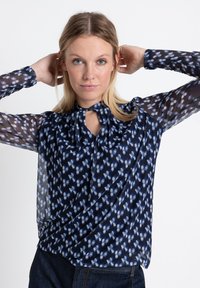 Marinebluse mit transparenten gemusterten Ärmeln, versehen mit einem Schlüsselloch-Ausschnitt und abstraktem blau-weißen Blumenmuster. Glattes Material, lockere Passform.