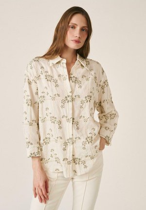 Mujer con una camisa abotonada de manga larga color beige claro con bordados florales y pantalones crema, de pie frente a un fondo neutro.