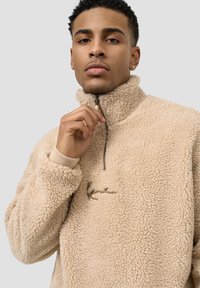 Beige fleece genser med høy krage, halv glidelås og broderte detaljer. Har ribbestrikkede mansjetter og en myk, teksturert overflate.